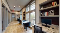 This single-floor business center and co-working space is thoughtfully designed to support a range of professional needs. The layout is defined by a long, open corridor lined with floor-to-ceiling windows along one wall, flooding the interior with abundant natural light and offering views of the surrounding outdoor landscape. Light wood-tone flooring runs the full length of the space, contributing to a clean and modern aesthetic.

Built-in granite-topped workstations are positioned along the window wall, each equipped with a desktop computer and ergonomic seating, providing dedicated spots for focused individual work. Interspersed between the workstations are small round tables paired with upholstered accent chairs in deep navy blue, offering casual seating options ideal for informal meetings or collaborative work sessions. Open shelving built into the walls displays books and decorative accessories, adding both function and visual warmth.

Along the opposite wall, a series of glass-paneled private rooms offer enclosed spaces for more focused or confidential work, each visible through full-height glass doors. Recessed lighting throughout ensures consistent illumination, complementing the natural light from the expansive windows. The overall ambiance is professional yet welcoming, balancing a refined, contemporary design with practical amenities suited for productivity.