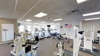 This single-floor fitness center offers a spacious and well-organized workout environment designed to accommodate a variety of exercise needs. The main exercise area is generously sized, featuring a row of treadmills and elliptical machines positioned to take advantage of the abundant natural light streaming in through multiple windows along the perimeter walls. The open floor plan allows for easy navigation between equipment stations, promoting a comfortable and efficient workout experience.

Strength training equipment, including cable machines and weight benches, is strategically arranged to maximize the use of available floor space without feeling overcrowded. The neutral-toned carpet flooring provides a practical surface throughout the facility. Overhead, a drop ceiling with recessed panel lighting ensures the space is consistently and evenly illuminated, complemented by a ceiling fan that aids in air circulation.

The walls are kept clean and minimal, with an American flag display adding a patriotic accent to the decor. A wall-mounted television is positioned for visibility from the cardio equipment area. The overall ambiance is bright, functional, and welcoming, making this fitness center a practical and inviting space for daily exercise routines.