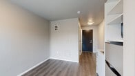 This studio unit at 6109 Roosevelt Way NE in Seattle's Roosevelt neighborhood offers a thoughtfully designed, efficient floor plan across a single level. The open-concept living area serves as the combined dining, kitchen, and living space, finished with durable wood-look flooring in a warm gray-brown tone and neutral light gray walls that create a clean, contemporary feel throughout.

The kitchenette is neatly integrated into the space, featuring white cabinetry with brushed hardware, a single-basin sink with a modern faucet, and a built-in shelving unit that houses a compact refrigerator and microwave — providing all the essential appliances in a space-efficient arrangement. A window at the end of the main living area allows natural light to filter in, contributing to an open and airy ambiance.

The entrance foyer leads directly into the main living area, defined by a dark-toned front door and a ceiling-mounted light fixture. A striking teal-painted ceiling accent in the transitional hallway adds a bold design detail that distinguishes the space.

The bathroom is well-appointed with a dark wood-toned vanity, a wall-mounted mirror with overhead lighting, a tall storage cabinet, and a full bathtub with shower — all finished in neutral, contemporary tones with vinyl flooring. Clean lines and functional design define every corner of this compact yet complete unit.