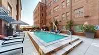 Located at 595 King Street in the heart of Charleston, South Carolina, this outdoor courtyard pool area offers a refined urban retreat set against the striking backdrop of a classic multi-story red brick building. The space is defined by its architectural character — large multi-pane windows, warm brick facades, and a U-shaped building envelope that creates a naturally sheltered and intimate atmosphere.

The centerpiece of the courtyard is a well-maintained rectangular swimming pool with vivid turquoise water, framed by clean tile coping and handrail entry points on both ends. The pool area is complemented by a spacious surrounding deck finished in light-toned stone pavers, offering ample room for lounging and socializing.

Furnishings are contemporary and thoughtfully arranged, featuring sleek lounge chairs and striped black-and-white umbrellas that evoke a classic resort aesthetic. Large modern planters filled with lush greenery and ornamental trees are positioned throughout the space, softening the urban setting with natural elements.

An adjacent outdoor terrace extends the usable area, equipped with additional seating arrangements including wicker lounge furniture with accent cushions, as well as an outdoor grilling station — ideal for gatherings. Open sky above allows for generous natural light throughout the day, making this courtyard a versatile and inviting outdoor amenity in the vibrant King Street corridor.