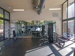 Located at 5155 S Old Peachtree Rd in Peachtree Corners, Georgia, this single-floor fitness center is designed to deliver a bright, open, and well-equipped workout environment. The space is defined by its dramatic floor-to-ceiling glass walls framed in sleek black metal, which flood the interior with abundant natural light and offer expansive views of the surrounding greenery and pool area. Soaring ceilings with exposed industrial-style ductwork add an urban, modern character to the space. The rubber-floored workout area is divided into distinct functional zones. The cardio section features a row of treadmills and elliptical machines positioned to take full advantage of the panoramic outdoor views, along with wall-mounted televisions for entertainment during workouts. The strength training area is equipped with a multi-station cable machine, adjustable benches, a full rack of dumbbells, and a dedicated free weights zone, all complemented by large wall-to-wall mirrors that visually expand the space and support proper form. Natural light streams across the dark rubber flooring throughout the day, creating an energizing atmosphere. The facility offers direct views of an outdoor pool, further enhancing the overall experience of this well-appointed community fitness center.