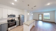 This thoughtfully designed one-bedroom, one-bathroom residence in Georgetown, Texas offers a clean, modern aesthetic across its single-floor layout. The open-concept kitchen and living area serves as the heart of the home, anchored by a granite-topped kitchen island with pendant lighting overhead. The kitchen is outfitted with white shaker-style upper cabinets, contrasting gray lower cabinets, a subway tile backsplash, stainless steel appliances including a refrigerator, range, microwave, and dishwasher, and a stainless steel sink. Light wood-tone flooring flows throughout the main living area, complementing the bright, neutral color palette. The living room features a glass exterior door and adjacent window that invite natural light and outdoor views. Built-in open shelving near the entry adds functional storage and visual interest. The bedroom offers plush carpeting, a ceiling fan, two windows for natural light, and direct access to both a walk-in closet with built-in shelving and the hallway leading to the bathroom. The bathroom is finished with a granite-topped gray vanity, a framed mirror, tile surround tub and shower combination, and light tile flooring, all accented with brushed nickel hardware throughout.