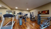 Located at 1425 NE Seventh Ave in Portland, Oregon, this fitness center offers a well-equipped, single-floor exercise facility designed to support a comprehensive workout routine. The space features warm hardwood-style flooring throughout, neutral beige walls accented with decorative artwork, and recessed lighting that creates a bright and welcoming environment.

The cardio section is outfitted with multiple treadmills, elliptical trainers, and a stationary bike, providing a variety of aerobic exercise options. A dedicated strength training area includes several weight machines for both upper and lower body workouts, along with a rack of dumbbells and stability balls for functional fitness exercises. A water cooler is conveniently positioned within the space to keep users hydrated during workouts.

Floor-to-ceiling mirrors line one wall, visually expanding the room and allowing users to monitor their form during exercise. A wall-mounted flat-screen television adds an entertainment element to cardio sessions. Large windows along one side of the room invite natural light and offer views of an outdoor courtyard or patio area, enhancing the overall ambiance. The well-organized layout ensures ample open floor space for movement between stations, making this fitness center a practical and comfortable amenity for regular use.
