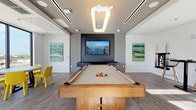 This single-floor game room is designed with a sleek, contemporary aesthetic that balances entertainment functionality with refined style. The centerpiece of the space is a full-size pool table constructed with a warm wood frame and neutral felt surface, positioned beneath a distinctive geometric pendant light that provides focused overhead illumination. Linear recessed lighting runs along the ceiling, complementing the pendant and ensuring consistent brightness throughout the room.

The walls are finished in a clean neutral palette, offering a crisp backdrop for two large abstract artworks featuring green and teal tones, which introduce color and visual interest to the space. A bold dark accent wall serves as a dramatic focal point, framing a large wall-mounted flat-screen television paired with a floating media console beneath it.

The flooring features wide-plank wood-look tiles in a cool gray tone, lending the space a modern, polished character while remaining durable for high-traffic use. A small seating area with vibrant yellow chairs is positioned to one side, adding a playful pop of color. The room benefits from natural light entering through windows along the perimeter, enhancing the bright, open atmosphere. Overall, the space is thoughtfully designed for recreation and social gathering.