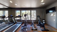 This single-floor fitness center offers a comprehensive workout environment designed to accommodate a variety of exercise routines. The cardio area is well-stocked with treadmills, an elliptical trainer, and stationary cycling bikes, each positioned to take full advantage of the large windows that line the walls. These expansive windows flood the space with natural light and frame views of an outdoor landscape featuring native desert trees and shrubs, creating an energizing atmosphere for exercise.

A floor-to-ceiling mirror runs along one wall, visually expanding the space and allowing users to monitor their form during workouts. The strength training section includes a multi-functional cable machine suited for a wide range of resistance exercises, as well as a dedicated free weight area with a full rack of rubber hex dumbbells organized in ascending weight increments. A wall-mounted television and ceiling fan add comfort and convenience to the environment.

The flooring features a distinctive patterned carpet tile design in tones of grey and amber, adding a modern aesthetic to the room. Neutral grey walls and recessed lighting contribute to the clean, contemporary feel of the space. A clearly posted fitness center policy board and sanitation station near the entrance reflect a commitment to cleanliness and user safety throughout.