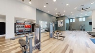 Located at 550 Webb Gin House Rd in Lawrenceville, GA, this single-floor fitness center is a spacious, thoughtfully designed facility built to support a full range of workout routines. The interior features light hardwood-style flooring throughout, soaring ceilings with clerestory windows that flood the space with natural light, and a clean, contemporary aesthetic defined by a crisp white and slate-blue color palette.

The facility is organized into distinct workout zones. A dedicated strength training area houses TKO cable machines, a Smith machine, a free-weight rack with a full dumbbell selection, and adjustable benches. Large wall mirrors enhance this section, creating a sense of openness while supporting proper form. An accessory station features organized shelving stocked with medicine balls, stability balls, and kettlebells for functional training.

The cardio zone is generously equipped with treadmills, stair climbers, ellipticals, and a WaterRower rowing machine. Expansive windows in this area offer views of an outdoor pool and courtyard, providing an energizing backdrop. A wall-mounted flat-screen TV adds entertainment for longer sessions.

Ceiling fans, recessed lighting, and double-door entry points contribute to the facility's airy, welcoming ambiance. The overall layout provides ample room to move freely between stations, making this fitness center a comprehensive and comfortable space for exercise.