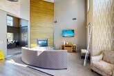 Located at 10209 Baltimore Ave in College Park, Maryland, this single-floor community clubhouse offers a thoughtfully designed layout across a generously sized footprint. The entrance sets an impressive tone with soaring double-height ceilings, bold gold-and-white wave-patterned wallcoverings, gleaming hardwood-style flooring, and oversized windows that flood the space with natural light. A striking sputnik chandelier anchors the grand entry hall, while a set of elegant black French doors provides a welcoming focal point.

The expansive living room is a standout feature, boasting dramatic double-height ceilings, a floor-to-ceiling horizontal slatted wood accent wall in warm golden tones, and a built-in fireplace. A curved sectional sofa, patterned area rug, and wall-mounted television complete the comfortable lounge setting.

The dedicated office suite serves as a functional business center, equipped with built-in cabinetry, workstation counters, a multifunction printer, and natural light from window blinds—ideal for productivity.

The galley-style kitchen is efficiently laid out with ample gray cabinetry, granite-style countertops, hardwood-style flooring, and black appliances including a refrigerator and dishwasher, providing full service convenience for events and gatherings.