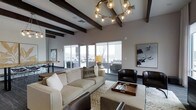 This thoughtfully designed single-floor space makes a strong architectural statement through its combination of elevated ceilings adorned with bold, dark-stained exposed wood beams that draw the eye upward and add warmth to the contemporary interior. A dramatic circular chandelier with globe-style Edison bulbs serves as a central focal point, casting a warm ambient glow throughout the main living area.

The living space is generously proportioned, centered around a plush, cream-toned sofa and complemented by rich, dark leather accent chairs that provide a striking contrast. A patterned area rug anchors the seating arrangement, while a wooden tray coffee table adds a refined, functional touch. Abstract wall art contributes to the sophisticated, modern aesthetic.

Large sliding glass windows and doors span the perimeter, inviting an abundance of natural light and offering views of the surrounding outdoor space. The seamless transition between the indoor seating area and the exterior terrace enhances the open, airy feel of the space. A secondary area in the background suggests additional functional zones, such as a dining or work area, further adding to the versatility of the layout. The overall ambiance is warm, modern, and welcoming.