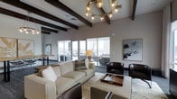 This thoughtfully designed single-floor space makes a strong architectural statement through its combination of elevated ceilings adorned with bold, dark-stained exposed wood beams that draw the eye upward and add warmth to the contemporary interior. A dramatic circular chandelier with globe-style Edison bulbs serves as a central focal point, casting a warm ambient glow throughout the main living area.

The living space is generously proportioned, centered around a plush, cream-toned sofa and complemented by rich, dark leather accent chairs that provide a striking contrast. A patterned area rug anchors the seating arrangement, while a wooden tray coffee table adds a refined, functional touch. Abstract wall art contributes to the sophisticated, modern aesthetic.

Large sliding glass windows and doors span the perimeter, inviting an abundance of natural light and offering views of the surrounding outdoor space. The seamless transition between the indoor seating area and the exterior terrace enhances the open, airy feel of the space. A secondary area in the background suggests additional functional zones, such as a dining or work area, further adding to the versatility of the layout. The overall ambiance is warm, modern, and welcoming.