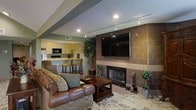 This single-floor space offers a thoughtfully designed open-concept layout that seamlessly connects the living and kitchen areas. The living room serves as the heart of the space, anchored by a striking floor-to-ceiling tiled fireplace that doubles as a media wall, complete with a large wall-mounted flat-screen television. Comfortable seating, including a classic leather sofa and upholstered armchairs, is arranged around a wooden coffee table atop a decorative area rug, creating a warm and inviting atmosphere. Recessed track lighting runs across the ceiling, providing flexible and modern illumination throughout the space. Lush potted plants add a natural, refreshing element to the décor. The kitchen is efficiently designed with light-toned cabinetry, black appliances, and a raised bar-height counter with bar stools, making it ideal for casual dining and entertaining. The open sightlines between the kitchen and living area enhance the sense of spaciousness and connectivity. Overall, the space strikes a balance between comfort and functionality, with warm earth tones, decorative accents, and practical finishes that create a cohesive and welcoming environment.