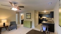 This single-floor space showcases an open-concept layout that seamlessly connects the living area, kitchen, and sleeping quarters. The living space features neutral beige walls and light-toned carpeting, creating a warm and inviting atmosphere. A ceiling fan with an integrated light fixture provides both airflow and ambient lighting overhead.

The kitchen is a standout feature, outfitted with dark espresso cabinetry that contrasts sharply against light tile flooring. Black appliances, including a refrigerator and range, are complemented by dark countertops and a raised breakfast bar with seating, making it functional for both cooking and casual dining.

A decorative green accent dresser acts as a visual centerpiece, bridging the living and kitchen areas while adding a pop of color. A framed nature-inspired artwork on the wall contributes to the space's understated aesthetic.

Visible through an open doorway, the en-suite bathroom continues the dark cabinetry theme, maintaining a cohesive design throughout. A thermostat on the wall and ceiling vents indicate individual climate control. Overall, the space is efficient and thoughtfully designed with a consistent, contemporary style.