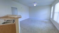This single-floor residence offers a clean and functional layout designed for comfortable everyday living. The main living area is finished with soft, plush wall-to-wall carpeting that adds warmth underfoot and contributes to the room's welcoming atmosphere. Neutral-toned walls and crisp white baseboards create a bright, airy feel throughout the space. A ceiling fan with an integrated light fixture is centrally mounted, providing both overhead illumination and effective air circulation year-round. The living area transitions naturally into the kitchen zone, which is defined by a raised bar-height counter finished in warm wood tones. This peninsula-style counter provides a convenient space for casual dining or meal prep and helps visually separate the kitchen from the living area without closing off the open-concept flow. The kitchen area also features a sink, adding practical utility to the space. Recessed outlets are thoughtfully positioned along the walls, and the overall design maintains a straightforward, uncluttered aesthetic. The single-floor layout ensures easy accessibility throughout, making this a practical and livable space suited to a range of occupants.