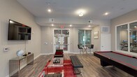 This single-floor amenity space is designed as a multifunctional community lounge, combining entertainment, relaxation, and social interaction in a cohesive, contemporary layout. The interior features durable wood-look flooring throughout, neutral gray and taupe wall tones, and recessed lighting that keeps the space well-illuminated.

The main lounge area is anchored by a wall-mounted flat-screen television, accompanied by sleek black lounge chairs with red accent pillows arranged around a glass coffee table atop a bold red area rug. A billiards table with a red felt surface serves as a central focal point, making the space ideal for social gatherings.

Adjacent to the lounge, a bistro-style high-top glass table with bar stools is positioned near large windows that overlook an outdoor pool area and the surrounding residential buildings, allowing natural light to flow freely into the space. A glass door provides direct access to this outdoor-facing area, enhancing the connection between interior and exterior.

Large interior glass windows offer views into a fitness center equipped with cardio machines, integrating the wellness facility visually within the amenity floor. A separate private office or meeting room with a glass-panel door is also accessible from the main space. The overall design balances functionality with modern aesthetics, creating an inviting environment for residents.