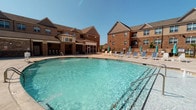 This amenity space combines an expansive outdoor pool area with a well-appointed indoor clubhouse, offering residents a comprehensive recreational retreat. The outdoor pool features clear, blue water set against a classic brick building backdrop, surrounded by a generous sun deck furnished with lounge chairs and teal-accented umbrellas. A covered outdoor bar area with high-top seating provides a shaded gathering spot for socializing.

The adjacent indoor clubhouse is designed with a contemporary aesthetic, featuring soaring vaulted ceilings that lend an open, airy feel throughout. The lounge area is anchored by a striking white stone fireplace wall with a mounted flat-screen television, complemented by plush teal seating arrangements and a patterned area rug over polished concrete floors. Large glass doors and windows allow abundant natural light to flow through the space.

A separate seating and café area showcases bold teal accent walls, mixed bar-height tables and chairs in teal and yellow upholstery, and a combination of pendant lighting and an ornate spherical chandelier. A coffee and refreshment station is conveniently accessible. Decorative artwork and sculptural accents throughout tie the cohesive, modern color palette together, creating a welcoming and functional communal environment.