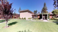 This single-story unit is part of a multi-building residential complex characterized by its warm stucco exteriors in earthy tones of terracotta and ochre, accented by brown-trimmed windows and sliding glass doors. The complex is thoughtfully arranged around expansive, well-kept communal lawns that create an open, park-like atmosphere, offering residents a generous shared outdoor environment beneath clear skies.

The grounds feature neatly maintained landscaping throughout, with a variety of shrubs, ornamental plants, and mature trees — including tall pines — lining paved walkways that connect the buildings. The greenery adds texture and shade, contributing to a pleasant and serene outdoor ambiance. A covered pergola area is visible near one of the buildings, providing a shaded outdoor gathering space.

The unit's entrance is ground-level, framed by trimmed hedges and accessed via a concrete pathway. Sliding windows flank the front door, and a wall-mounted air conditioning unit is installed above the entryway. The unit benefits from direct access to the surrounding green spaces without the need to navigate stairs, distinguishing it from upper-floor units within the complex. The overall setting conveys a calm, well-established community environment with an emphasis on outdoor livability.