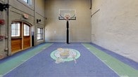 This impressive indoor basketball court offers a striking combination of athletic functionality and architectural character. The court surface is finished in a blue and green modular tile system, prominently featuring a Boston Celtics logo at center court, complete with painted boundary lines and arc markings. A regulation-height basketball hoop with a clear backboard stands centered at one end, ready for full-court play.

The space is enclosed by textured, plastered walls that rise to exceptional heights, lending the court an open, airy feel. Natural light filters in through tall transom windows positioned high along the walls, as well as through windows flanking the court's entrance. The soaring ceiling height accommodates full gameplay and adds to the grand atmosphere of the space.

At one end of the court, two arched wooden double doors with wire-glass panels serve as the primary entry points, framed by exposed brick surrounds that hint at the building's historic character. Utility conduits and fire safety equipment are neatly integrated along the walls. The overall ambiance blends the energy of an active athletic facility with the warmth of a well-preserved, architecturally distinctive structure — making this basketball court as visually engaging as it is functional.