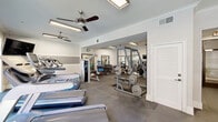 Located at 925 S Main St in Grapevine, TX, this fitness center offers a comprehensive workout environment spread across a single floor. The space is divided into two distinct zones, each purpose-built to support a full range of exercise routines.

The cardio area is bathed in natural light through large floor-to-ceiling glass doors and windows that overlook an outdoor pool and courtyard. Multiple treadmills and stationary bikes are positioned to take advantage of these views, while ceiling fans provide added ventilation and comfort. Motivational wall decals reinforce the energetic atmosphere of the space.

The strength training area is equally well-appointed, featuring a multi-station cable machine, power racks loaded with weight plates, a full dumbbell rack, kettlebells, medicine balls, exercise mats, and stability balls. Large wall-mounted mirrors throughout both zones enhance the sense of space and allow users to monitor their form during workouts.

The interior finishes are clean and modern, with neutral gray-toned walls accented by horizontal striping, crown molding, and durable large-format tile flooring suited for heavy use. Overhead fluorescent lighting supplements natural light in the strength training zone. The overall design is functional yet polished, creating an inviting and well-maintained facility for residents of the Grapevine community.