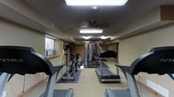 This fitness center is a dedicated, single-floor exercise facility designed to support a comprehensive workout routine. The room is laid out in a long, open floor plan that comfortably accommodates multiple pieces of equipment without feeling cramped. Cardio machines, including treadmills and an elliptical trainer, are arranged along the central and peripheral areas of the room, providing ample space for users to move freely between stations. A multi-station strength training machine anchors the middle of the space, offering a range of resistance-based exercises. The flooring consists of a combination of carpet and protective rubber mats beneath the equipment, ensuring both comfort and safety. Overhead, a ceiling fan provides air circulation, complementing the fluorescent lighting that keeps the space well-lit throughout. A skylight panel further enhances the brightness of the room. Natural light filters in through windows positioned along one wall, adding to the open and energizing atmosphere. The neutral, warm-toned walls and low-profile ceiling contribute to a functional and focused environment well-suited for fitness activities. The overall ambiance is clean, practical, and purposefully arranged to serve users looking for a well-rounded exercise experience.