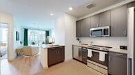 Located at 10 Commerce Dr in New Rochelle, NY, this thoughtfully designed single-floor residence offers a seamless blend of modern aesthetics and functional living. The open-concept layout connects the kitchen and living area, creating a generous, airy feel throughout.

The kitchen is a standout feature, outfitted with dark charcoal cabinetry, light quartz countertops, a classic white subway tile backsplash, and a full suite of stainless steel appliances including a range, microwave, and dishwasher. A central kitchen island with an integrated sink provides additional prep space and serves as a natural dividing point between the kitchen and living area.

The living area is bright and welcoming, with large windows dressed in emerald green curtains that invite natural light while adding a touch of color to the otherwise neutral palette. Light hardwood flooring flows consistently throughout the main living spaces, enhancing the sense of openness.

The bedroom is a serene retreat, featuring large windows with outdoor views, soft neutral tones, and a spacious walk-in closet with built-in shelving and hanging space. The bathroom is clean and contemporary, finished with large-format floor tiles and smooth linear wall tiles surrounding a combination tub and shower, complemented by crisp white fixtures throughout.