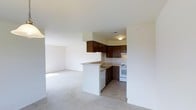 This single-floor space presents an open-concept design that seamlessly connects the kitchen and living area, creating a cohesive and functional layout. The kitchen is outfitted with dark wood cabinetry that extends to the upper walls, providing generous storage capacity. A peninsula counter serves as a natural divider between the kitchen and the living space, offering additional prep area and casual seating potential. The kitchen includes a white electric range and a dishwasher integrated beneath the counter, alongside a sink conveniently positioned on the peninsula. Light-toned countertops complement the darker cabinetry, and the flooring transitions from tile in the kitchen to carpet in the living area. The living space is notably open and bright, with neutral white walls that enhance the sense of airiness. Flush-mount ceiling lighting provides even illumination throughout. The overall layout is practical and efficient, with clean lines and a neutral color palette that offers a versatile base for a variety of interior styles.
