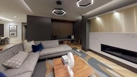 This elegantly designed single-floor amenity space blends contemporary aesthetics with warm, inviting materials to create a refined communal environment. The main lounge area is anchored by a sleek fireplace set within a clean, stone-clad surround, complemented by light hardwood flooring laid in a striking herringbone pattern and an artfully patterned area rug. Seating is thoughtfully arranged, featuring a generous sectional sofa with blue accent pillows, a leather lounge chair with a brass frame, and additional accent seating — all centered around a solid wood coffee table. Dramatic hexagonal pendant lights hang from the dark ceiling, casting a modern, atmospheric glow throughout the space. A vertical wood slat partition serves as an architectural focal point, framing a cozy built-in bench nook behind. Along one wall, a well-appointed refreshment station offers dark cabinetry, a coffee machine, under-counter refrigeration, and a wall-mounted display screen beneath a layered wood slat ceiling detail. Wide hallways connect the various areas, lined with curated framed artwork, and lead to a warm wood-paneled private room with its own bench seating and display panels. The overall ambiance is polished and contemporary, balancing dark, moody tones with bright, light-filled accents.