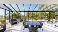 This thoughtfully designed single-floor office space blends modern professionalism with an inviting, biophilic aesthetic. The centerpiece of the space is a stunning glass solarium enclosed by floor-to-ceiling steel-framed windows and a sloped glass roof, flooding the interior with natural light while offering panoramic views of surrounding trees and the cityscape beyond. The solarium features tiled flooring, a round dining table with woven chairs, and an array of lush tropical plants, creating a greenhouse-like retreat ideal for informal gatherings or focused work.

The open main workspace is finished with neutral carpeting and white walls, anchored by a bold sculptural chandelier overhead. Built-in shelving with cabinetry, a cushioned banquette, and curated artwork add warmth and character to the collaborative area. Exposed concrete columns lend an industrial contrast to the otherwise refined interior.

Private conference rooms are enclosed by sleek glass-panel partitions with dark wood-framed doors, offering visual transparency while maintaining acoustic separation. Additional enclosed rooms, accessible via keycard entry, provide space for focused work or smaller meetings.

Throughout the space, an abundance of potted plants — from towering monstera to banana palms — reinforces a vibrant, nature-forward atmosphere that distinguishes this workspace as both functional and inspiring.