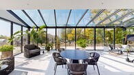 This thoughtfully designed single-floor office space blends modern professionalism with an inviting, biophilic aesthetic. The centerpiece of the space is a stunning glass solarium enclosed by floor-to-ceiling steel-framed windows and a sloped glass roof, flooding the interior with natural light while offering panoramic views of surrounding trees and the cityscape beyond. The solarium features tiled flooring, a round dining table with woven chairs, and an array of lush tropical plants, creating a greenhouse-like retreat ideal for informal gatherings or focused work.

The open main workspace is finished with neutral carpeting and white walls, anchored by a bold sculptural chandelier overhead. Built-in shelving with cabinetry, a cushioned banquette, and curated artwork add warmth and character to the collaborative area. Exposed concrete columns lend an industrial contrast to the otherwise refined interior.

Private conference rooms are enclosed by sleek glass-panel partitions with dark wood-framed doors, offering visual transparency while maintaining acoustic separation. Additional enclosed rooms, accessible via keycard entry, provide space for focused work or smaller meetings.

Throughout the space, an abundance of potted plants — from towering monstera to banana palms — reinforces a vibrant, nature-forward atmosphere that distinguishes this workspace as both functional and inspiring.