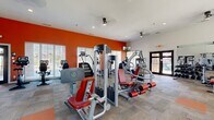 This fitness center offers a spacious, single-floor layout designed to accommodate a comprehensive workout experience. The facility is entered through a set of sleek, glass-paneled double doors, which provide a clear sightline into the workout area and create a welcoming, transparent entry point. Adjacent to the entrance is a small waiting area furnished with a slatted bench and a beverage cooler, along with posted fitness rules for member reference.

Inside, the main workout floor is defined by bold, burnt-orange accent walls that energize the space, contrasting sharply with the clean white ceiling above. The flooring consists of a patterned, multi-tone carpet with orange tile accents, providing both comfort and visual interest underfoot.

Natural light filters in through multiple large windows fitted with horizontal blinds, giving the space a bright and airy atmosphere. Wall-mounted televisions are positioned throughout for entertainment during workouts.

The equipment selection is extensive and well-organized. Cardio offerings include a row of Life Fitness treadmills, elliptical trainers, stair climbers, and stationary bikes. Strength training is supported by a variety of selectorized cable machines, leg press and extension equipment, and a dedicated free weights area featuring a full dumbbell rack, weight bench, and large wall mirrors. Ceiling fans and recessed lighting ensure a comfortable environment throughout.