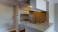 This single-floor space offers a practical and well-organized layout designed for comfortable everyday living. The kitchen serves as a central feature, thoughtfully arranged to maximize efficiency without sacrificing usability. A peninsula countertop with a granite-style surface extends into the space, creating a natural divide between the kitchen and the adjoining area while offering additional workspace and casual seating potential. White upper cabinetry provides generous storage, complemented by a continuous countertop that houses a stainless steel sink and a smooth electric cooktop. White appliances, including a full-size refrigerator and dishwasher, contribute to the kitchen's clean, cohesive aesthetic. A range hood overhead ensures proper ventilation during cooking. The walls feature a neutral taupe tone that lends warmth and versatility to the space, while wood-style flooring grounds the interior with a touch of texture. A fire extinguisher is conveniently mounted near the kitchen entry, reflecting a practical attention to safety. Overall, the space presents a straightforward, move-in-ready environment with a functional kitchen as its defining highlight.