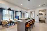 Located at 700 Avett Ln in Charlotte, NC, this thoughtfully designed single-floor amenity space offers a well-rounded environment for both relaxation and productivity. The open-concept living area serves as the heart of the space, featuring large-format tile flooring, recessed lighting, and an abundance of natural light streaming through floor-to-ceiling windows dressed with sheer blue curtains. A central elongated bar-height island with a wood-grain finish and leather-upholstered bar stools anchors the room, complemented by a wall-mounted flat-screen TV. Varied seating arrangements — including a plush lounge sofa, a bold blue high-back booth, and small café-style tables — create distinct zones for socializing or quiet work. A tray ceiling with a circular pendant light adds architectural interest and depth to the main area, while barn-style sliding doors introduce a stylish design detail. The private office features a built-in wood desk, carpeted flooring, and curated wall art, offering a quiet retreat for focused work. A dedicated laundry room rounds out the functionality of this versatile space, making it a comprehensive and polished community amenity.