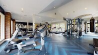 This spacious fitness center is thoughtfully designed to support a full range of workout routines, combining a clean, modern aesthetic with motivational character. The flooring throughout is a standout feature — a stylish herringbone-patterned tile in cool blue-grey tones that grounds the space with a polished yet dynamic feel.

The facility is organized into distinct workout zones. A dedicated cardio area lines the perimeter near large floor-to-ceiling windows, housing multiple treadmills, ellipticals, and stationary bikes that allow users to exercise with views of the surrounding greenery. The strength training section is equipped with cable machines, a multi-functional Smith machine, free weights, and benches to accommodate a variety of resistance exercises.

A separate functional training area includes stability balls, medicine balls, resistance bands, and a mat board, complemented by a full-length mirror and an arched window that adds architectural charm. Exposed whitewashed brick walls and rustic brick columns contrast beautifully with the bright white ceilings and recessed lighting. A bold motivational mural reading "A Champion — Live Like It" anchors one end of the space, while decorative oversized boxing gloves add personality to a vaulted alcove. Ceiling fans and a wall-mounted TV complete the well-rounded, energizing environment.