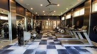 This single-level fitness center is designed with both functionality and style in mind. The space is anchored by a striking black-and-white checkered floor that adds a bold visual contrast to the dark, sophisticated wall treatment throughout. Recessed lighting is distributed evenly across the ceiling, ensuring the entire area is well-illuminated, while two ceiling fans provide consistent air circulation during workouts.

The cardio zone, positioned along the right side of the room, features a row of Matrix treadmills and elliptical machines, each equipped with individual display consoles. On the opposite side, a selection of Matrix strength and cable machines is arranged along a full-length mirrored wall, which enhances the sense of space and allows users to monitor their form. Additional equipment, including a rowing machine, rounds out the strength-training offerings.

Natural light enters through a set of windows along one wall and glass-panel double doors at the far end, which also provide a view of greenery outside. The overall ambiance is sleek and modern, creating an environment well-suited for focused, effective training sessions.