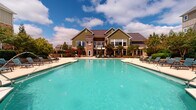 Located at 1000 Antietam Creek Dr in Fayetteville, NC, this outdoor amenity space is thoughtfully designed to deliver a resort-like experience. At its centerpiece is a generously sized, freeform swimming pool with crystal-clear turquoise water, surrounded by a broad concrete sundeck lined with aqua-toned lounge chairs — ideal for relaxing under the Carolina sun.

The pool area is framed by mature trees, manicured shrubs, and carefully maintained landscaping beds that provide both shade and a sense of natural privacy. A stained wood pergola anchors an outdoor dining and grilling area, offering a shaded retreat with seating for small gatherings. Additional Adirondack-style chairs are positioned throughout the deck for casual lounging.

The main building features a striking combination of sage green siding and stacked stone masonry accents, complemented by a standing-seam metal roof. Large windows and glass doors open directly onto the pool deck, seamlessly connecting interior and exterior spaces. Controlled access to the pool area is provided via a wrought iron gate with stone pillar surrounds, ensuring a secure and well-maintained environment. The overall atmosphere is clean, welcoming, and well-appointed — designed to serve as a premier outdoor gathering destination for the community.