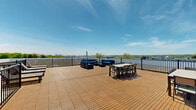 Located at 6515 Ridge Avenue in the Roxborough neighborhood of Philadelphia, this rooftop deck is a spacious outdoor amenity designed for both relaxation and social gatherings. The deck features warm-toned composite wood tile flooring that spans a generously sized open-air platform, bordered by sleek dark metal railings that ensure safety while preserving unobstructed sightlines.

The space is thoughtfully arranged into distinct functional zones. A lounge area is outfitted with plush blue cushioned seating, ideal for casual conversation and unwinding, while multiple outdoor dining tables with chairs accommodate group meals and entertaining. A set of reclining sun loungers provides a dedicated area for sunbathing, positioned to take full advantage of the open sky above.

A blue-sided utility structure anchors one section of the deck, housing mechanical equipment and providing access points to the rooftop. Adjacent to the main deck, a green roof section adds an eco-conscious design element to the building.

The most striking feature of this rooftop is its sweeping panoramic views — stretching across the treetops and waterway to the Philadelphia skyline in the distance. Under open skies, the deck offers an elevated outdoor retreat with an open, airy ambiance that makes it a standout communal space.