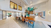 This thoughtfully designed amenity space delivers a dynamic blend of social and relaxation zones across a single, expansive floor. Polished terrazzo floors flow throughout, unifying each area while reflecting abundant natural light that pours in through floor-to-ceiling black-framed windows and glass entry doors. The centerpiece of the main gathering area is a striking, large-scale tropical botanical mural that spans an entire wall, creating a vivid visual anchor for the double-height dining zone below. A long communal wooden dining table surrounded by an eclectic mix of chairs invites gathering, while pendant globe lights cascade overhead to add warmth. Adjacent to this, a bright game lounge features a full-size shuffleboard table and suspended swing chairs, delivering a playful, resort-inspired atmosphere. A dedicated lounge area offers plush sofas, a round sculptural coffee table, and a mounted display screen, complemented by sliding glass-panel doors that allow for flexible privacy. A second lounge zone incorporates hanging greenery, additional soft seating, and private enclosed rooms with glass walls for intimate meetings or quiet use. A built-in cabinetry wall with open shelving, curated art, and a tile backsplash defines the reception corridor, completing this well-appointed communal environment.