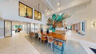 This thoughtfully designed amenity space delivers a dynamic blend of social and relaxation zones across a single, expansive floor. Polished terrazzo floors flow throughout, unifying each area while reflecting abundant natural light that pours in through floor-to-ceiling black-framed windows and glass entry doors. The centerpiece of the main gathering area is a striking, large-scale tropical botanical mural that spans an entire wall, creating a vivid visual anchor for the double-height dining zone below. A long communal wooden dining table surrounded by an eclectic mix of chairs invites gathering, while pendant globe lights cascade overhead to add warmth. Adjacent to this, a bright game lounge features a full-size shuffleboard table and suspended swing chairs, delivering a playful, resort-inspired atmosphere. A dedicated lounge area offers plush sofas, a round sculptural coffee table, and a mounted display screen, complemented by sliding glass-panel doors that allow for flexible privacy. A second lounge zone incorporates hanging greenery, additional soft seating, and private enclosed rooms with glass walls for intimate meetings or quiet use. A built-in cabinetry wall with open shelving, curated art, and a tile backsplash defines the reception corridor, completing this well-appointed communal environment.