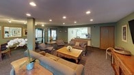 This single-floor space is thoughtfully designed to support communal living and social interaction across several distinct areas. The main lounge is a spacious, open area finished with sage green walls, plush carpeting, and recessed lighting that creates a warm, inviting ambiance. Comfortable sofas and armchairs are arranged around a decorative coffee table, while a wall-mounted flat-screen television and a media console serve as focal points. A sliding glass door leads to an outdoor patio area, allowing natural light to fill the room. An adjacent seating nook features a small dining table with upholstered chairs, track lighting overhead, and framed artwork on the walls, adding a refined touch. A pass-through opening connects this area to a compact, well-equipped kitchen outfitted with white cabinetry, light countertops, a double stainless steel sink, and small appliances including a toaster oven. The kitchen is practical and organized, with ample cabinet storage. Throughout the space, sage green walls and carpeted flooring create a cohesive, calming aesthetic, while the varied lighting solutions ensure each area is well-illuminated and functional for a range of activities.