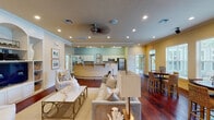 This single-story club house is thoughtfully designed to serve as a premier gathering space for its community. Rich hardwood flooring flows throughout, unifying the various social areas under warm, neutral-toned walls that create an inviting and relaxed atmosphere.

The main lounge area is a standout, boasting soaring coffered ceilings with recessed lighting that enhances the sense of grandeur. A classic white-mantled fireplace anchors one wall, flanked by large windows that flood the room with natural light and offer views of the outdoor pool area. Comfortable seating arrangements make this an ideal space for both casual relaxation and organized events.

A secondary lounge area features a built-in entertainment center with shelving, display niches, and a television, complemented by decorative millwork and columned architectural details that add a refined, residential feel.

The kitchen is well-appointed with white cabinetry, a mosaic tile backsplash, granite countertops, stainless steel appliances, and a curved breakfast bar — fully equipped to support catered events or community functions.

A dedicated dining area with a round table and wicker seating sits adjacent to a bank of windows overlooking the pool, offering a bright and casual spot for meals or conversation. The space also includes access to additional rooms, likely for administrative or meeting purposes, rounding out this versatile and elegantly appointed club house.