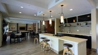 This thoughtfully designed single-floor space combines modern aesthetics with functional amenities, creating an inviting environment for gathering and socializing. The open-concept layout seamlessly connects a fully equipped kitchen with expansive communal dining and lounge areas, all unified by warm wood-look flooring that runs throughout.

The kitchen area is a standout feature, anchored by a sleek, light-toned island with a built-in gas cooktop and bar-height seating for several adjustable industrial-style stools. Pendant lighting with rectangular frosted glass shades hang above the island, providing both task lighting and a striking design element. Dark cabinetry lines the perimeter, complemented by a neutral tile backsplash, open shelving, and a wall-mounted television.

Beyond the kitchen, the space opens into a spacious lounge and dining area, furnished with dark-toned tables and chairs arranged to accommodate groups of various sizes. Recessed lighting illuminates the space from above, while decorative framed artwork and tall potted plants add warmth and life to the surroundings. Architectural details, including tiered ceiling treatments and rich accent walls in deep wood tones, lend depth and character to the overall design, resulting in a polished yet welcoming communal atmosphere.