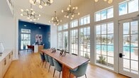 Located at 111 Patterson Ln in Meridianville, Alabama, this single-floor dining room is a well-appointed, light-filled space designed with a contemporary aesthetic. The room's most striking architectural feature is its soaring ceiling height, which amplifies the sense of openness and grandeur. An entire wall of floor-to-ceiling gridded windows and a glass-paned door floods the interior with abundant natural light while framing direct views of an outdoor pool area, creating a seamless visual connection between indoors and outdoors.

The dining area is anchored by a substantial live-edge walnut dining table surrounded by teal upholstered chairs, complemented overhead by multiple mid-century modern sputnik chandeliers with globe bulbs that serve as decorative focal points. Light oak hardwood flooring runs throughout, adding warmth and continuity to the space.

Along one wall, built-in cabinetry with light wood and brass hardware provides functional storage beneath a clean white countertop. A curated gallery wall of colorful artwork adds visual interest. An arched doorway leads into an adjacent alcove finished in deep navy blue paneling, housing a compact desk workspace accented by bold artwork and pendant lighting. A hallway off the main room provides access to a restroom finished with navy subway tile and mosaic flooring.