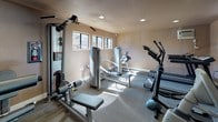 This fitness center is a single-floor facility thoughtfully designed to support a full range of exercise routines. The space is finished with warm neutral-toned walls and durable carpet flooring suited for a high-traffic workout environment. Recessed lighting throughout ensures consistent, even illumination, while multiple horizontal windows along the walls invite natural light and offer views of the surrounding outdoor area.

The room is outfitted with an extensive array of equipment to accommodate both cardio and strength training needs. Cardio machines include treadmills, an elliptical trainer, and a stationary upright bike, providing a variety of aerobic workout options. For strength training, the space features multiple cable and resistance machines, including a multi-function cable station and dedicated press and fly machines, offering targeted muscle group exercises.

A wall-mounted air conditioning unit and ceiling ventilation ensure climate comfort during workouts. A water cooler is conveniently stationed within the room, and a posted fitness center rules board keeps the facility organized. A glass-paneled entry door provides easy access and connects the space to the surrounding outdoor walkway area. The overall ambiance is clean, functional, and well-maintained, making it a practical destination for daily fitness routines.