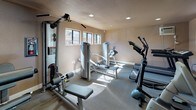 This fitness center is a single-floor facility thoughtfully designed to support a full range of exercise routines. The space is finished with warm neutral-toned walls and durable carpet flooring suited for a high-traffic workout environment. Recessed lighting throughout ensures consistent, even illumination, while multiple horizontal windows along the walls invite natural light and offer views of the surrounding outdoor area.

The room is outfitted with an extensive array of equipment to accommodate both cardio and strength training needs. Cardio machines include treadmills, an elliptical trainer, and a stationary upright bike, providing a variety of aerobic workout options. For strength training, the space features multiple cable and resistance machines, including a multi-function cable station and dedicated press and fly machines, offering targeted muscle group exercises.

A wall-mounted air conditioning unit and ceiling ventilation ensure climate comfort during workouts. A water cooler is conveniently stationed within the room, and a posted fitness center rules board keeps the facility organized. A glass-paneled entry door provides easy access and connects the space to the surrounding outdoor walkway area. The overall ambiance is clean, functional, and well-maintained, making it a practical destination for daily fitness routines.