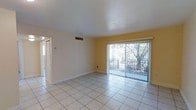 This single-story space features a straightforward, functional layout centered around a bright and open living area. The room is finished with large, light-toned ceramic floor tiles that extend throughout, lending a clean and cohesive look to the interior. The walls are painted in a soft yellow hue, adding a subtle warmth to the otherwise neutral palette, while white baseboards provide a crisp contrast along the perimeter.

A prominent sliding glass door dominates one wall of the living area, fitted with vertical blinds that allow for adjustable light control. The door opens to an outdoor area where mature trees and a fenced yard are visible, offering a connection to the exterior environment and allowing natural light to filter into the room.

The ceiling is smooth and flat, with a centrally located HVAC vent indicating a ducted climate control system. A wall-mounted thermostat is also visible near the entryway, providing convenient temperature management. An adjacent hallway or entry corridor connects the living space to the remainder of the unit, suggesting additional rooms beyond the main living area. Overall, the space presents a clean, uncluttered environment well-suited for practical everyday use.