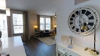 This well-appointed single-floor space offers a cohesive and stylish living environment defined by rich, dark hardwood flooring that flows seamlessly throughout. The living area serves as the centerpiece of the space, anchored by a classic dark sofa, a patterned area rug, and warm accent lighting from matching table lamps that create an inviting ambiance. Large windows dressed with elegant gold-toned drapes allow generous amounts of natural light to pour in, offering views of the surrounding urban environment. A ceiling fan overhead adds both function and a subtle design touch to the room. The layout transitions smoothly into a secondary area, where a white desk or vanity is accompanied by a decorative oversized Roman numeral wall clock, adding a bold, artistic statement to the space. Thoughtful styling details — including brass accents, layered textiles, and carefully chosen décor — lend the space a warm, curated feel. The overall aesthetic balances contemporary and classic elements, resulting in a space that feels both functional and visually engaging, ideal for comfortable everyday living.