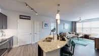 This thoughtfully designed single-floor residence combines contemporary style with functional living across a well-organized open layout. The kitchen is a standout feature, showcasing dark espresso cabinetry, granite countertops, a white subway tile backsplash, stainless steel appliances including a refrigerator, range, and built-in microwave, as well as a wine refrigerator. A curved granite-topped island with an undermount sink doubles as a breakfast bar with seating, seamlessly connecting the kitchen to the dining and living areas. The living room is bright and inviting, with soft blue-gray accent walls, wood-look plank flooring, large windows, and a sliding glass door that opens to a private balcony. Ceiling fans are present in both the living room and bedrooms. The primary bedroom offers plush carpeting, generous natural light from multiple windows, and direct access to an en-suite bathroom featuring a walk-in glass shower, quartz countertop vanity, and dark cabinetry. The second bedroom also features carpeted flooring, an accent wall, a ceiling fan, and a walk-in closet. A second full bathroom includes a soaking tub with tiled surround and a matching dark vanity. Track lighting and pendant fixtures add to the modern aesthetic throughout the home.