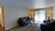 This single-floor space features a living room designed for comfort and functionality. The room is finished with wall-to-wall carpeting in a neutral tone, complementing the light-colored walls that contribute to a bright and open atmosphere. A large sliding glass door dominates one wall, providing direct access to an outdoor area while flooding the space with natural light and offering views of greenery outside. Vertical blinds accompany the sliding door, allowing for adjustable light control and privacy. The living area comfortably accommodates a dark leather sofa accented with patterned throw pillows, paired with a round glass-top coffee table supported by a dark wood frame. A floor lamp positioned near the sliding door provides warm ambient lighting during evening hours. The clean, simple layout of the room allows for flexible furniture arrangement. Smooth, flat ceilings and clean baseboards throughout contribute to the room's tidy, well-maintained appearance. The overall ambiance is modest and functional, with natural light and outdoor views serving as notable highlights of the space.