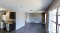 This single-floor space presents a straightforward, functional layout centered around an open living area. The room is finished with dark hardwood-style flooring that runs throughout, offering a rich contrast against the neutral gray walls and crisp white baseboards and ceiling. A flush-mount ceiling light fixture provides overhead illumination, while a window along one wall allows natural light to brighten the interior.

The living area is generously sized, offering an open and unobstructed floor plan that provides flexibility for various furniture arrangements. A doorway visible toward the rear of the space leads to an adjoining room, suggesting a connected layout that flows naturally between areas. A wall-mounted HVAC vent near the ceiling ensures climate control throughout.

To one side, a partial view of the kitchen reveals dark cabinetry and a stone-style countertop, hinting at a practical and updated kitchen space. The overall aesthetic is clean and modern, with a neutral color palette that creates a calm, versatile environment. The combination of dark flooring and gray tones gives the space a contemporary feel, well-suited for a variety of interior styles.