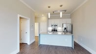 This single-floor space showcases a well-designed open-concept kitchen that serves as a central focal point. The kitchen is outfitted with a full suite of stainless steel appliances, including a French door refrigerator with an in-door ice and water dispenser, a stainless range with multiple burners, and a built-in microwave. Crisp white cabinetry extends to the ceiling, providing generous storage, while a large island topped with speckled granite countertops offers additional workspace and casual seating potential. Two sleek pendant lights hang above the island, complementing the recessed lighting and a flush-mounted ceiling vent for a cohesive, modern aesthetic. Crown molding runs along the ceiling perimeter, adding a refined architectural detail throughout. The rich, dark hardwood-style flooring contrasts elegantly with the light walls and white cabinetry, grounding the space with warmth and depth. Neutral wall tones create a versatile backdrop, while the open layout allows for a natural flow between the kitchen and surrounding living areas. Doorways visible from the kitchen hint at additional private rooms beyond.