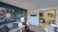 This single-floor space offers a thoughtfully designed open-concept layout that seamlessly connects the living area to the kitchen. The living room immediately draws attention with a bold teal accent wall that adds depth and character to the space, complementing the neutral white walls and light-toned hardwood-style flooring that runs throughout. The room is well-proportioned, providing comfortable space for seating arrangements and everyday living.

A framed coastal landscape artwork anchors the main wall, reinforcing a calm, contemporary aesthetic. Recessed transitions between rooms are clean and well-finished, with grey-toned door frames adding a subtle modern touch.

Visible through the open passage, the kitchen features dark cabinetry with a clean, modern profile, light countertops, and stainless steel appliances including a dishwasher. The kitchen maintains the same light hardwood-style flooring, creating visual continuity throughout the space.

Overall, the space presents a cohesive, modern design with a balanced mix of bold color choices and neutral tones, creating an inviting and functional environment suited for comfortable day-to-day living.