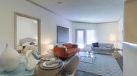 This single-floor residence offers a thoughtfully designed open-concept layout that connects the dining and living areas into one cohesive, comfortable space. The living room is a focal point of the home, featuring a bold rust-orange tufted sofa paired with a sleek light gray couch, anchored by a geometric-patterned area rug and a minimalist glass coffee table. A striking abstract wall art piece adds a contemporary touch, complementing the clean, neutral color palette throughout. Large windows allow generous natural light to fill the space, creating a bright and airy atmosphere. Crown molding along the ceiling adds a refined architectural detail, while the textured ceiling contributes subtle depth. The dining area is positioned adjacent to the living room, featuring a round dark-toned table set with neatly arranged place settings, ideal for entertaining. A glimpse into the bedroom reveals a well-appointed sleeping area with consistent styling. The overall ambiance is modern and inviting, balancing functional design with curated décor elements that give the space a polished, livable character throughout.